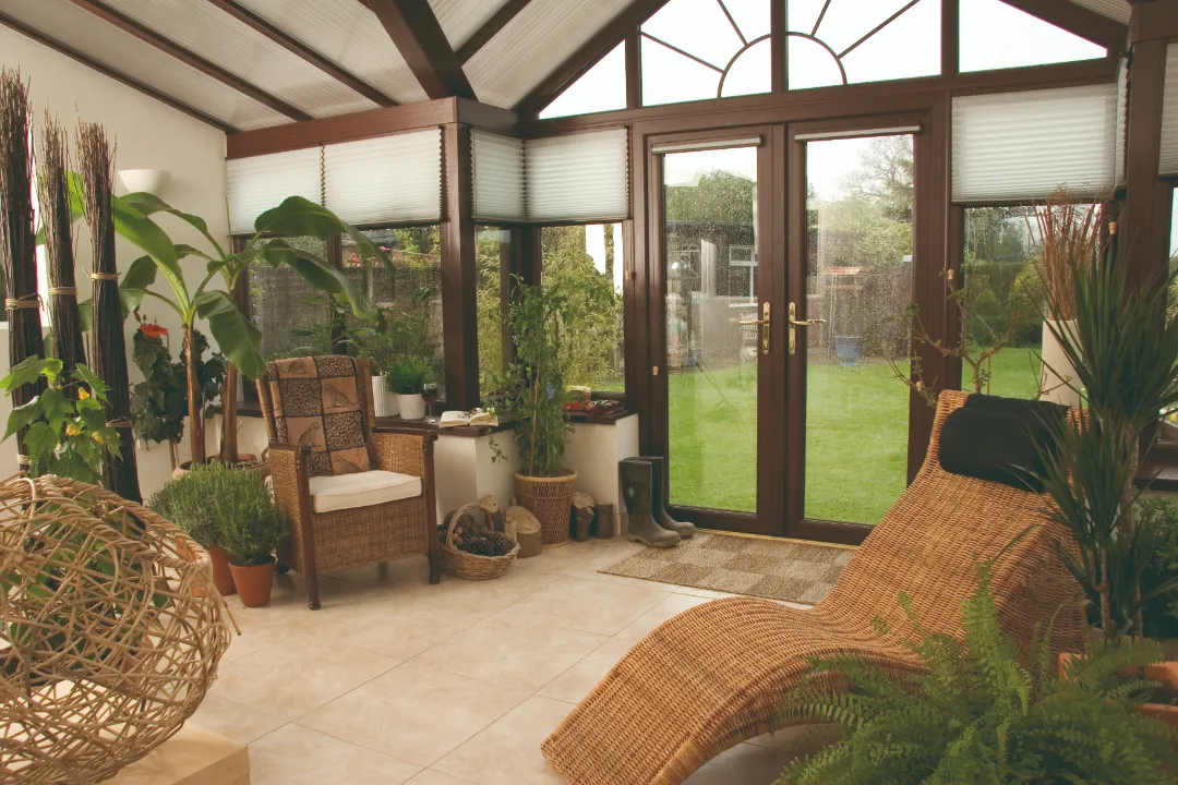 Interior of a dark wood-framed sunroom filled with house plants, featuring a wicker lounge chair, a armchair, and French doors looking out into a green lawn.