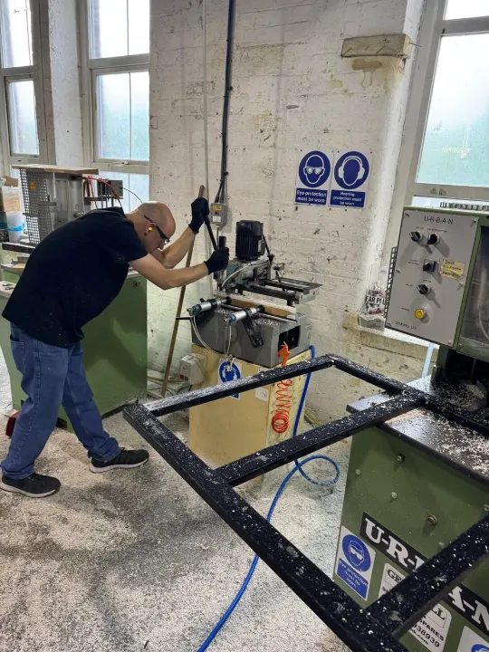 A worker wearing protective gear operating machinery inside a window frame manufacturing workshop.