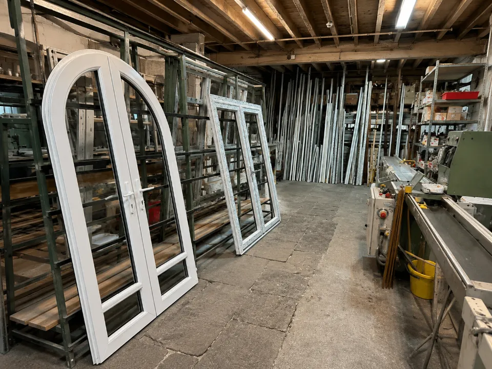 Finished white arched French doors and window frames stacked inside a factory workshop.