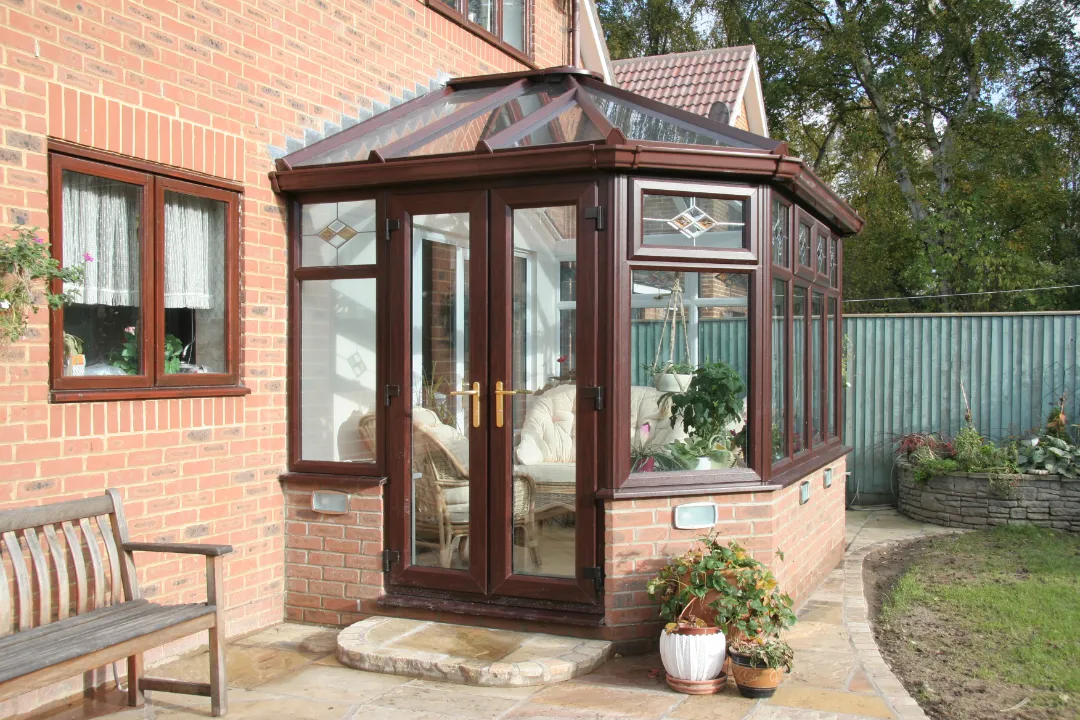 A dark wood-grain conservatory featuring a glass roof, leaded glass top windows, and double French doors attached to a red brick house.