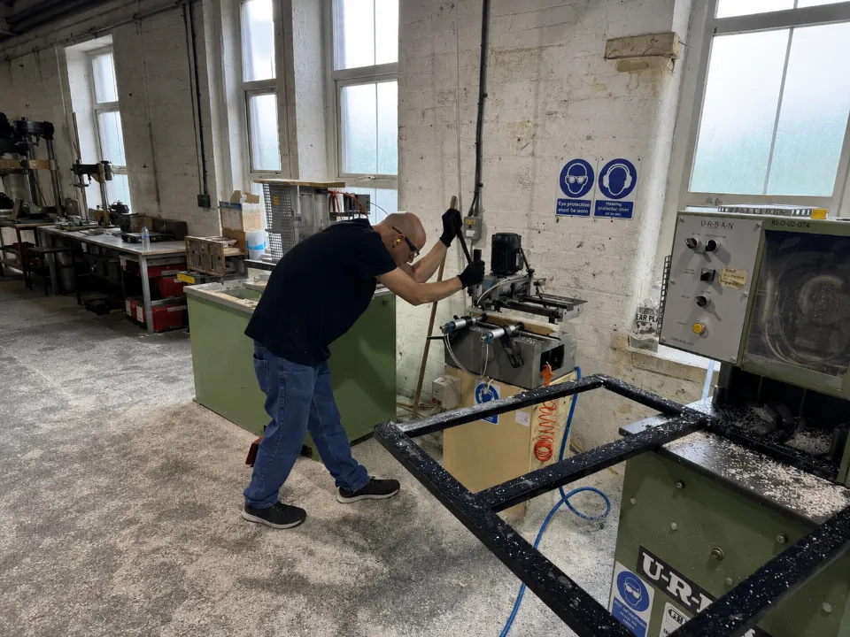 A worker wearing protective gear operating machinery inside a window frame manufacturing workshop.
