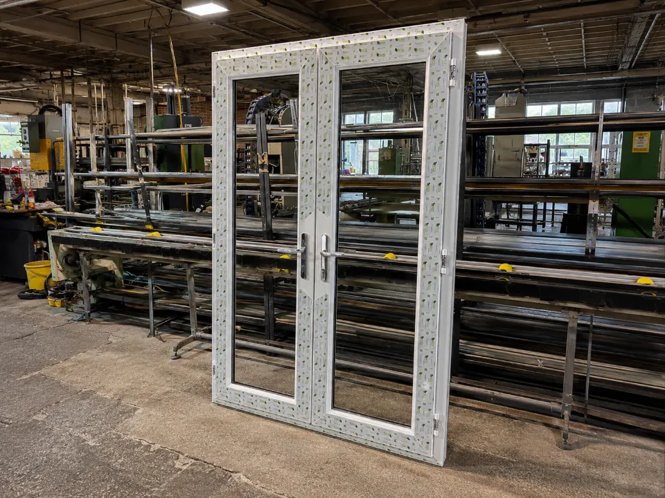 A set of white UPVC French double doors with protective film standing on a factory floor.