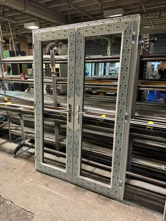 A set of white UPVC French double doors with protective film standing on a factory floor.