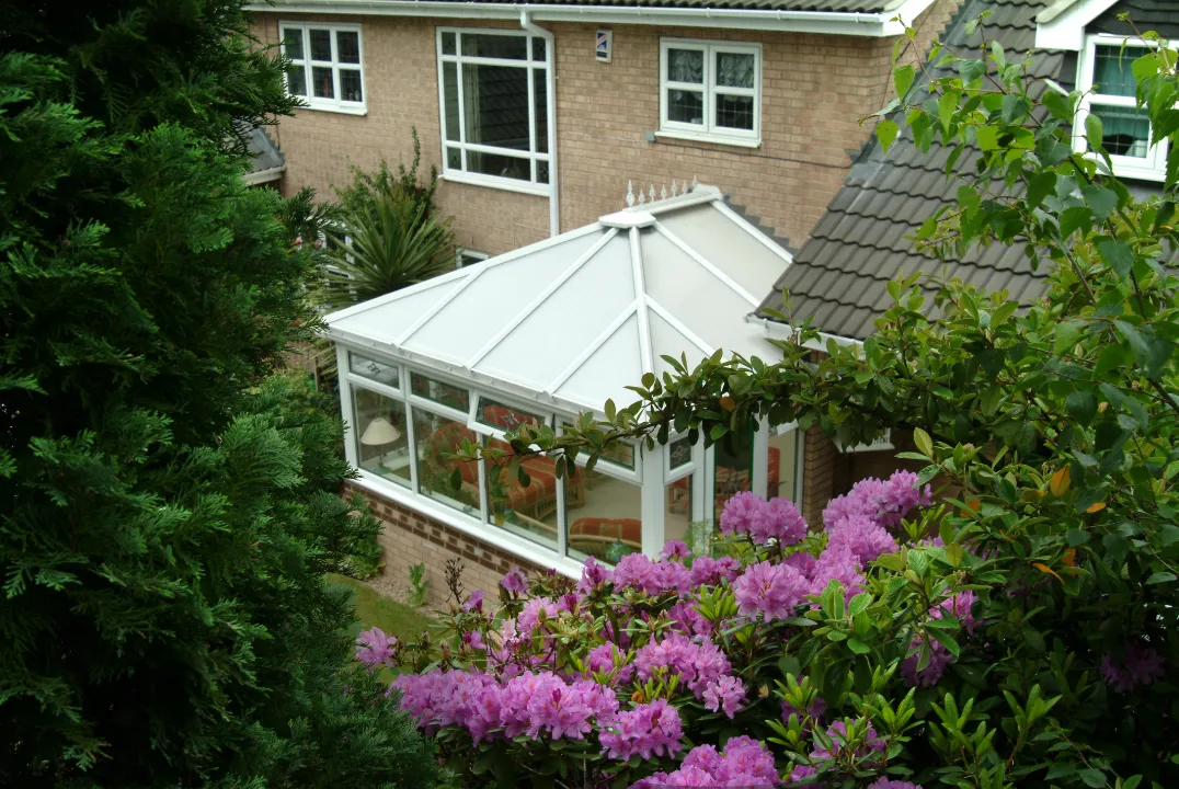 Elevated view of a white Victorian-style conservatory extension nestled among lush trees and vibrant pink rhododendron flowers.