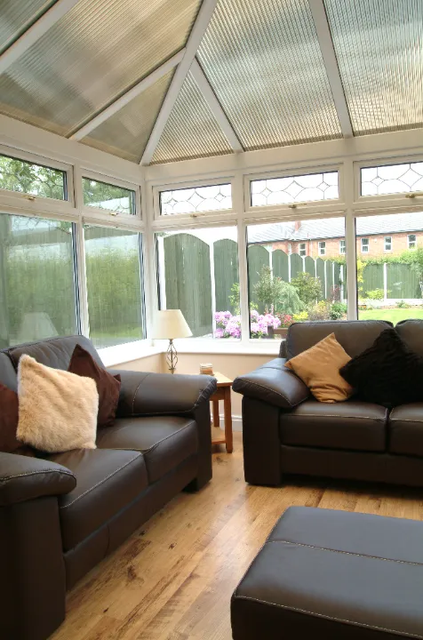 Interior view of a cozy sunroom furnished with dark leather sofas, light wood flooring, and pitched roof panels looking out onto a garden.