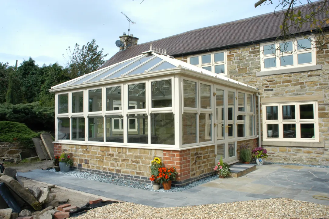 Off-white conservatory with large windows built on a stone base, attached to a traditional stone house.