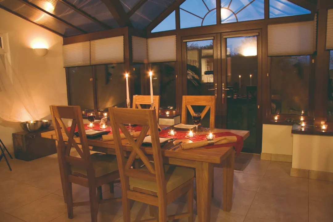 Interior view of a conservatory dining room set for a candlelit evening dinner with a wooden table and chairs under warm lighting.