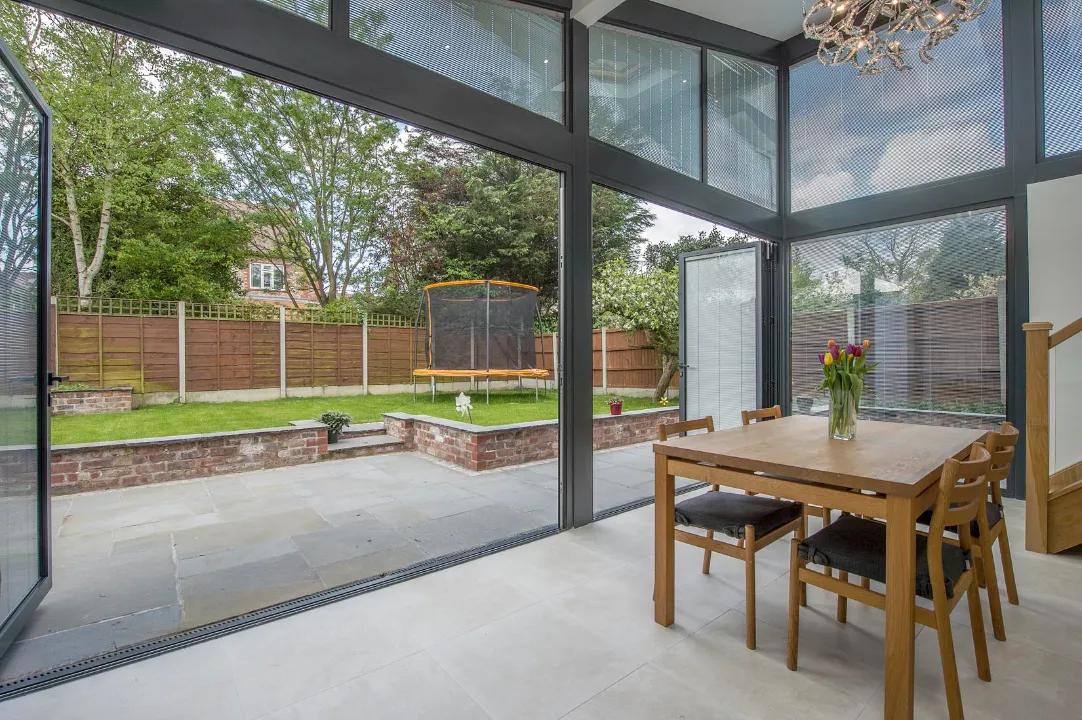 Open dark-framed bi-fold glass doors creating a seamless transition between an indoor dining area and a stone patio back garden with a trampoline.
