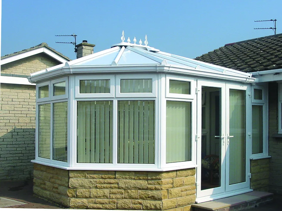 Exterior view of a white uPVC conservatory built on a stone base, featuring integrated blinds and French doors leading outdoors.