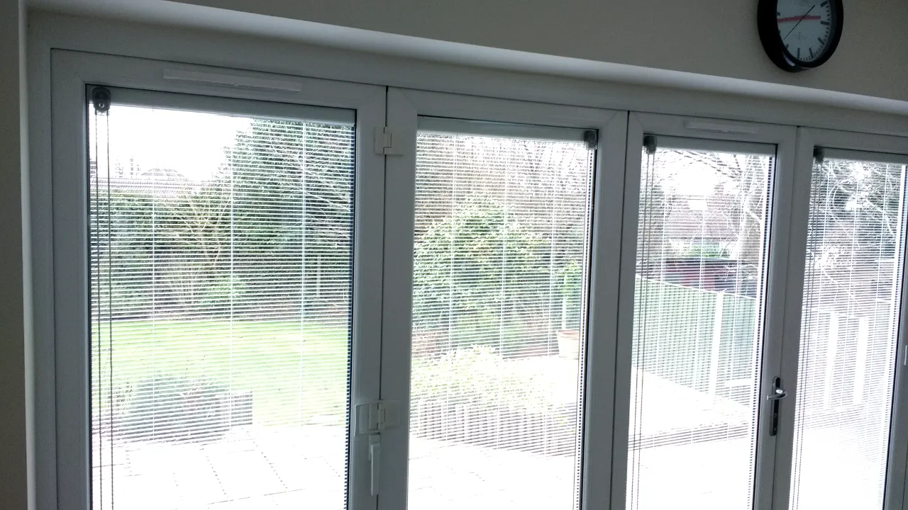 view from inside a room looking out white-framed bi-fold glass patio doors with integrated blinds opening to a green lawn.