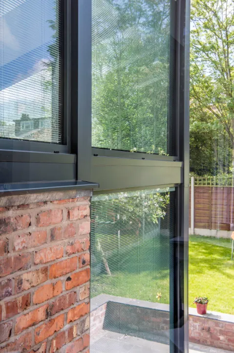 Close-up interior view of tall black-framed window panels featuring integrated blinds next to an exposed red brick wall.