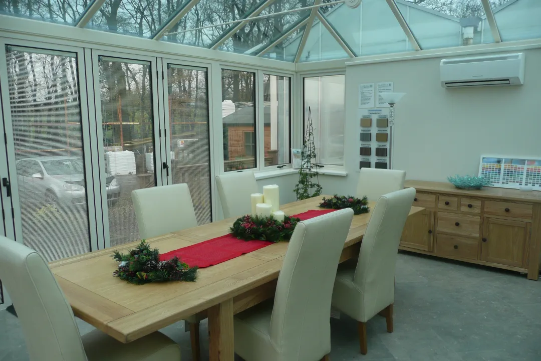 A large wooden dining table with white chairs set inside a bright glass-roof conservatory with white bifold doors and integrated window blinds.