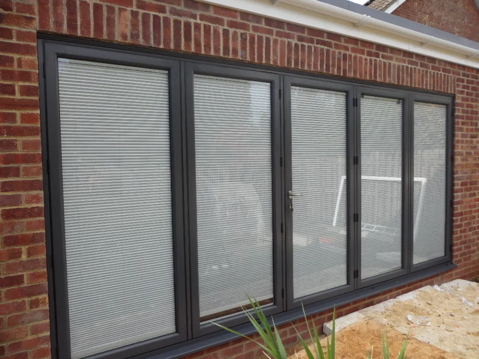 Exterior view of dark grey framed bi-fold glass doors with internal blinds installed on a red brick home extension.