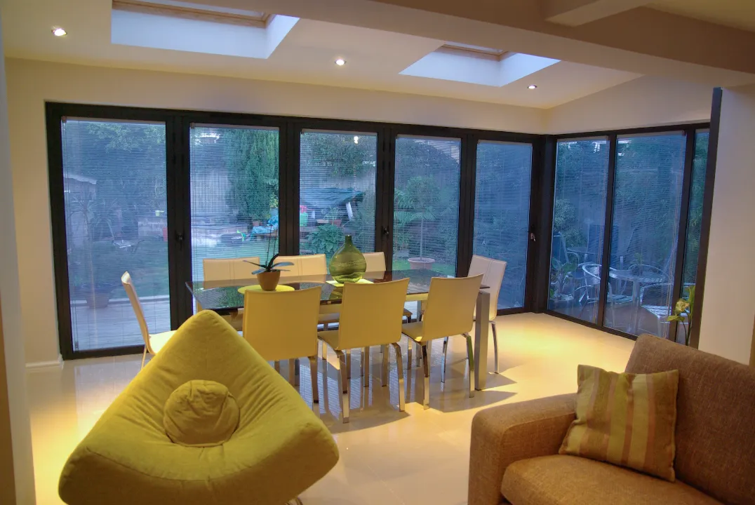 A modern dining and living space featuring floor-to-ceiling dark frame glass doors with built-in blinds overlooking a backyard garden.