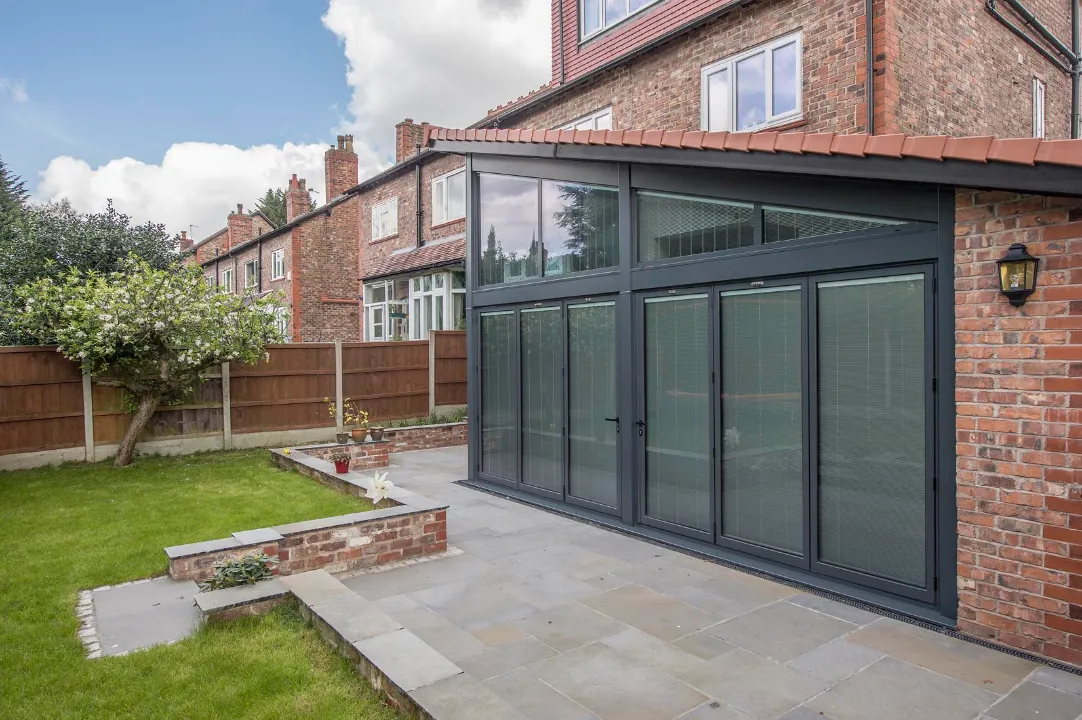 Exterior view of a modern brick home extension featuring dark gray bi-fold glass doors with integrated blinds leading out to a stone patio and grassy garden.