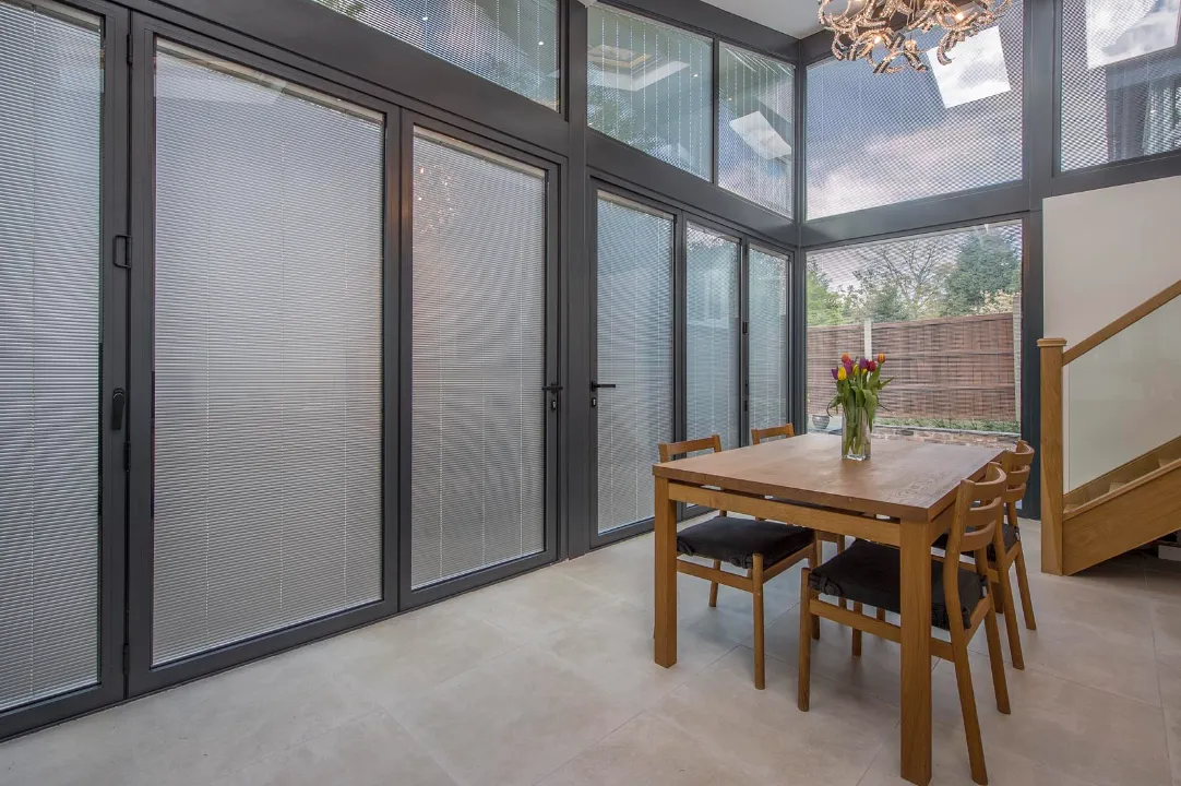 Interior view of a bright dining space showcasing floor-to-ceiling glass wall panels and bi-fold doors equipped with integrated blinds.