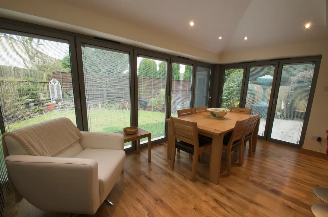 A bright home extension featuring a wooden dining table, a white armchair, and surrounding glass patio doors with built-in blinds.
