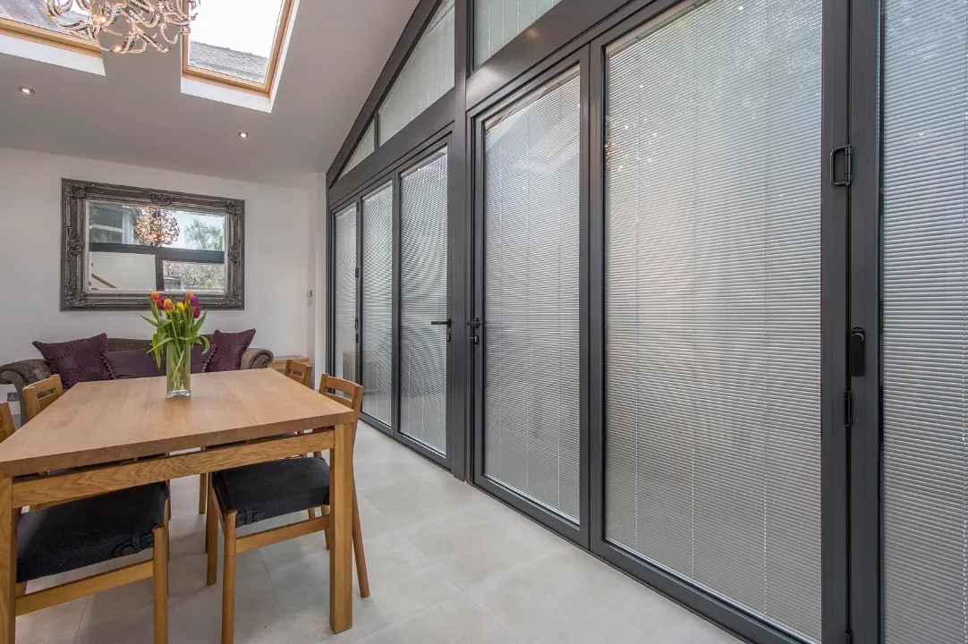 An interior dining room with a wooden table and chairs positioned in front of tall dark-framed glass doors fitted with integrated blinds.