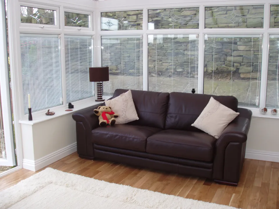 A dark brown leather couch with two white accent pillows and a small teddy bear set inside a sunroom featuring white window frames with built-in blinds.