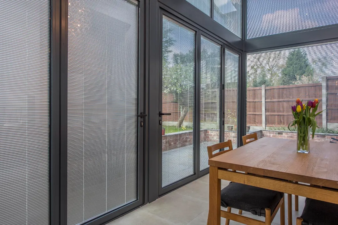 A wooden dining table with a vase of colorful tulips placed near large dark-framed glass doors fitted with integrated blinds.
