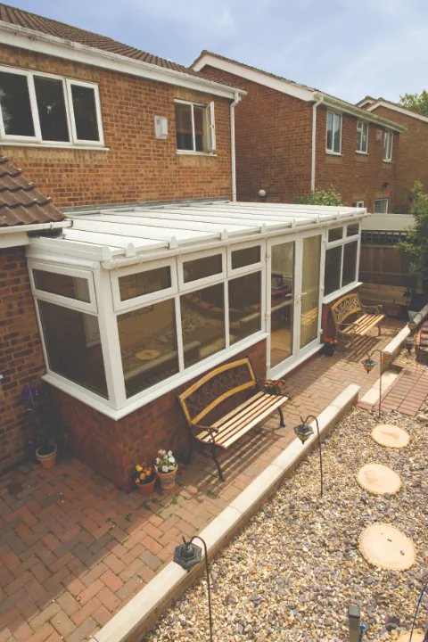 High-angle view of a white lean-to conservatory extension built onto a red brick house with a paved patio, gravel garden, and wooden benches.