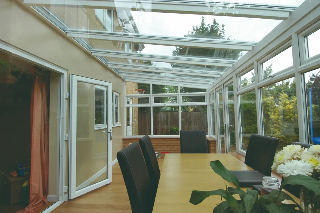 Interior view of a lean-to sunroom with a clear glass roof, light wood flooring, and a long wooden dining table surrounded by black chairs.