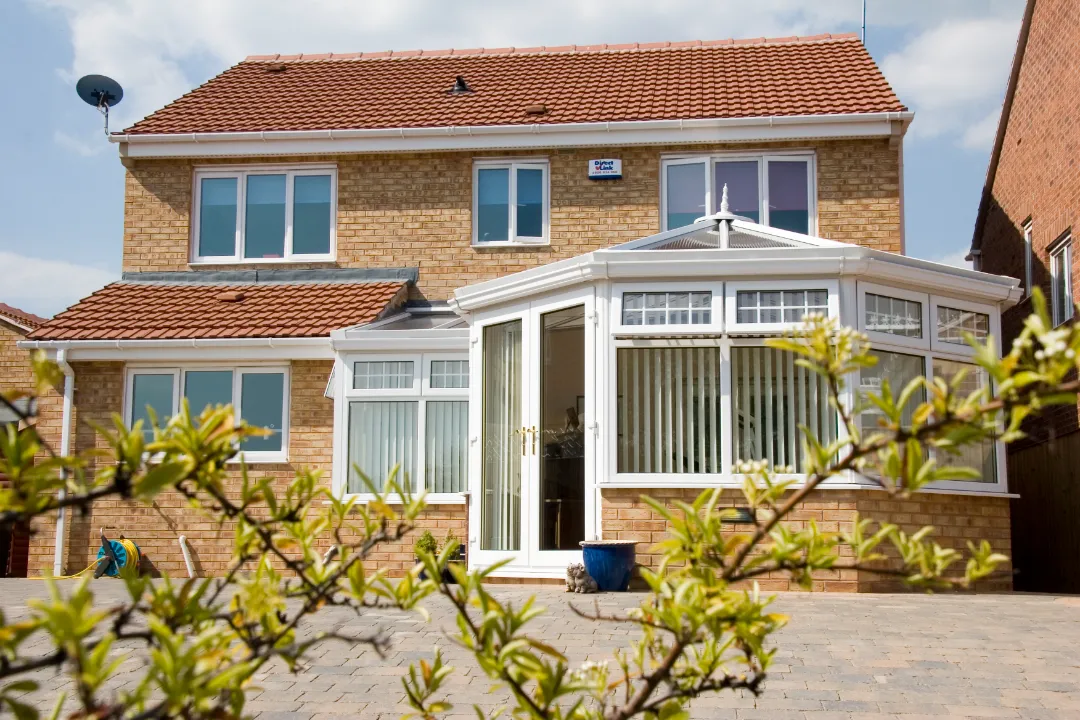 Large white conservatory with vertical blinds attached to a two-story brick house, framed by green tree branches in the foreground.