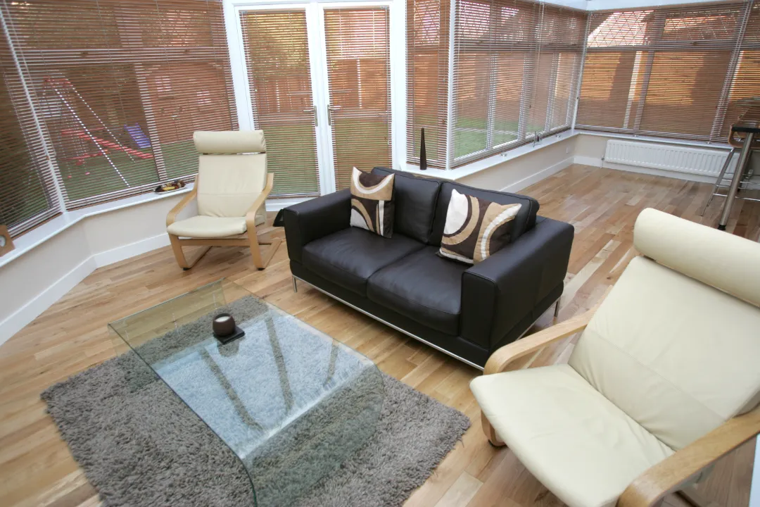 Interior of a modern sunroom with light wood flooring, horizontal window blinds, a dark leather sofa, and cream armchairs around a glass coffee table.