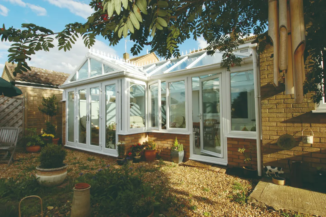 White glass conservatory built onto a brick base set in a gravel garden surrounded by potted plants.