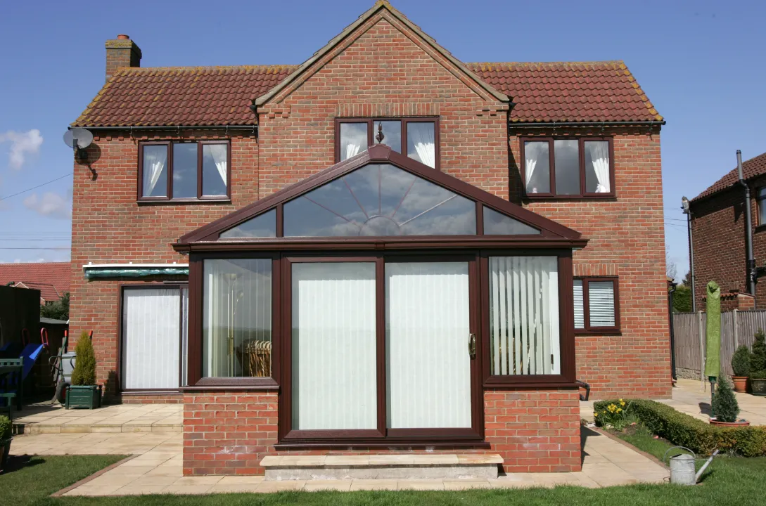 Direct rear view of a two-story red brick house with a central dark wood-grain conservatory featuring sliding glass patio doors and a sunburst transom window.