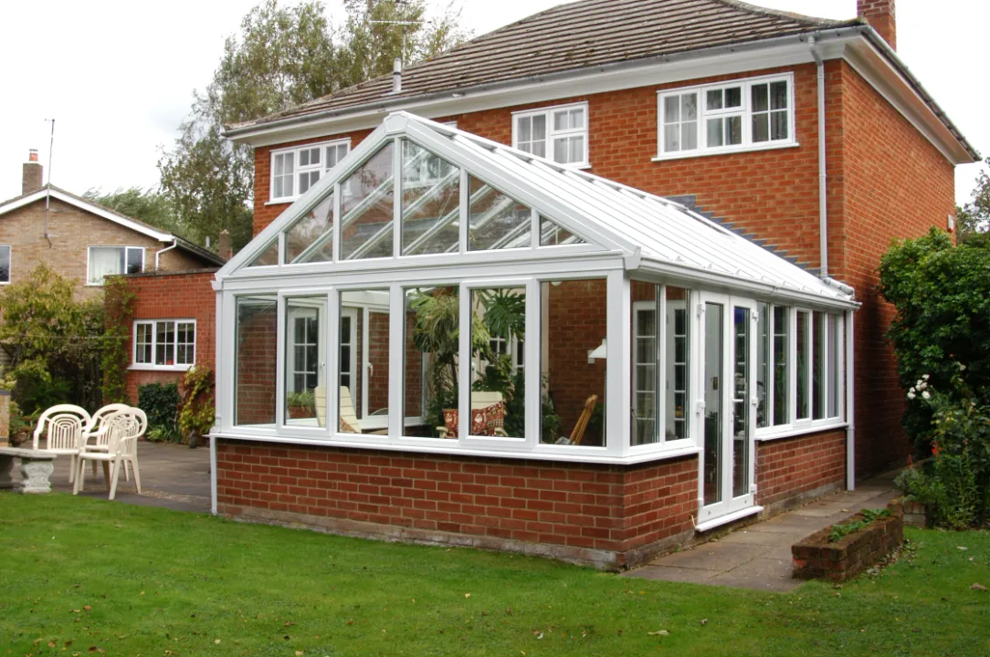 Large white conservatory with a high glass pitched roof and brick base attached to the back of a red brick two-story home.