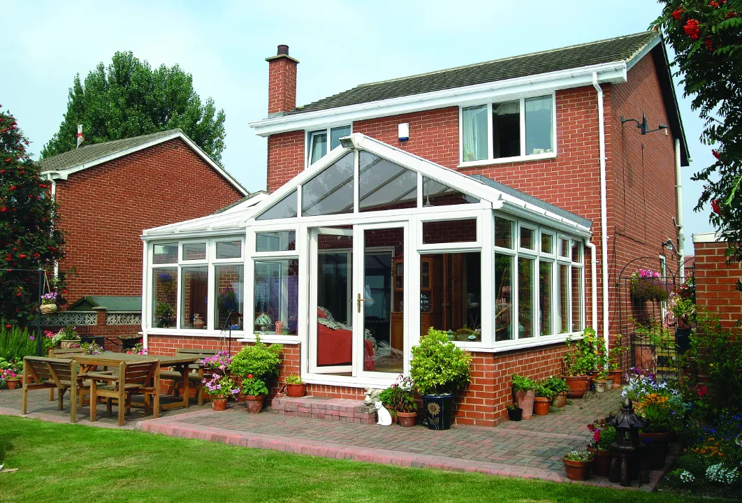 White gable-front conservatory extension attached to the back of a two-story red brick home with a paved patio area.