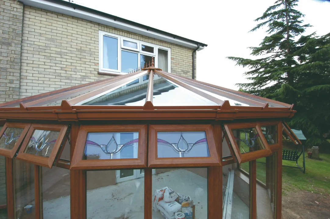 Close-up of a wood-grain conservatory roof structure showing opening top transom windows with decorative stained glass panels.