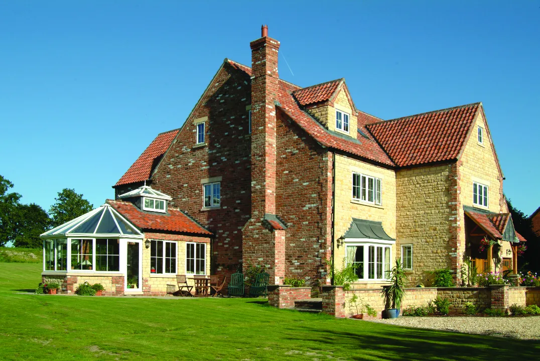 A large two-story brick and stone country home featuring a white conservatory extension on the side, surrounded by a spacious green lawn.
