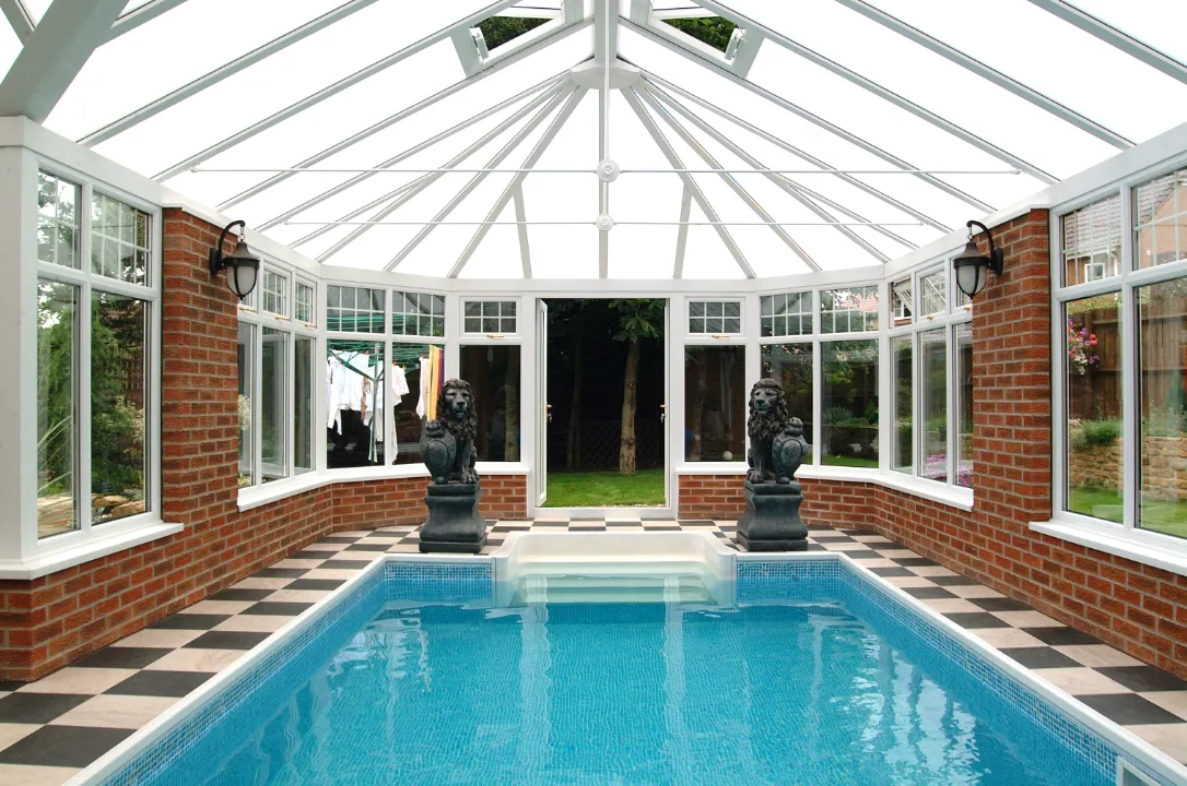 Interior view of a conservatory enclosing an indoor swimming pool, framed by brick walls, stone lion statues, and a glass pitched roof.