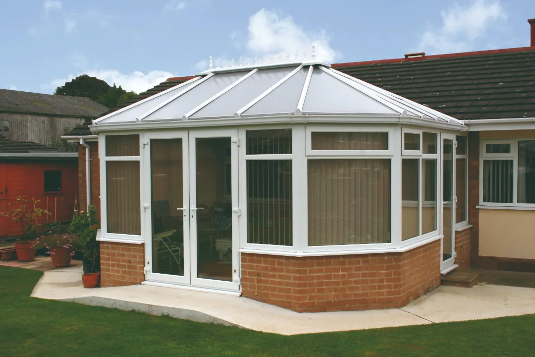 Exterior view of a white uPVC conservatory built on a low brick wall, featuring French doors and vertical window blinds.