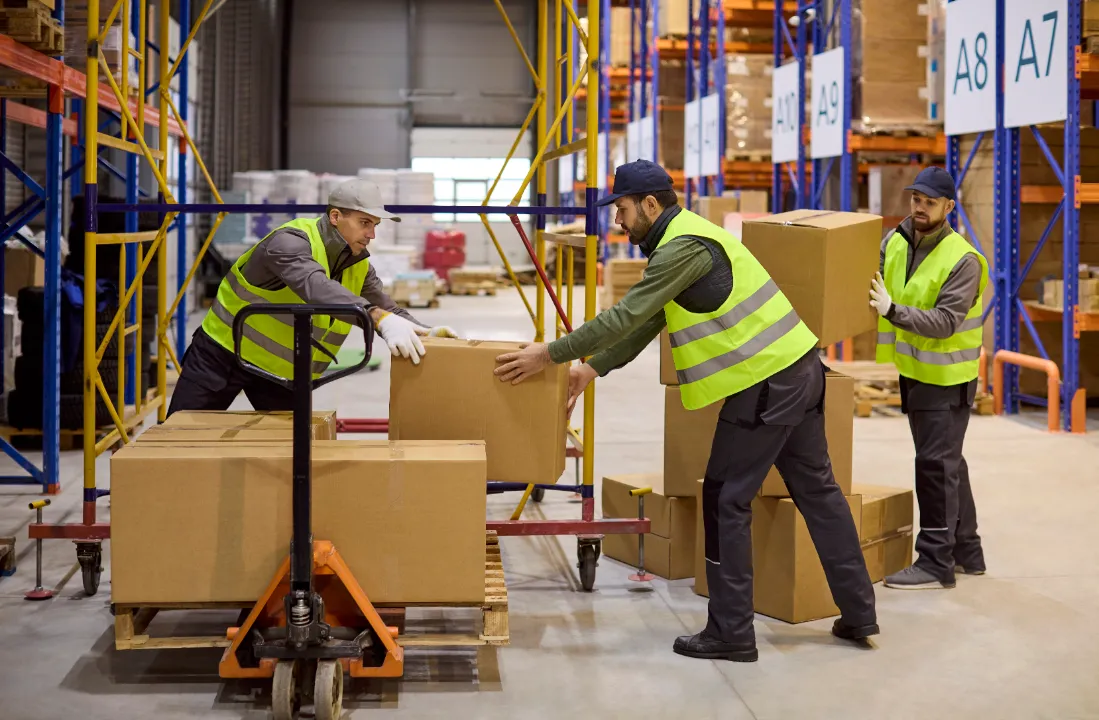 Warehouse workers wearing high-visibility vests loading cardboard boxes onto a hydraulic pallet jack.
