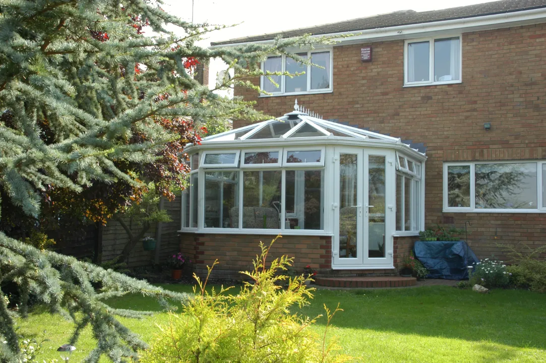 Exterior view of a white uPVC Victorian-style conservatory attached to a two-story red brick house with a green lawn and trees.