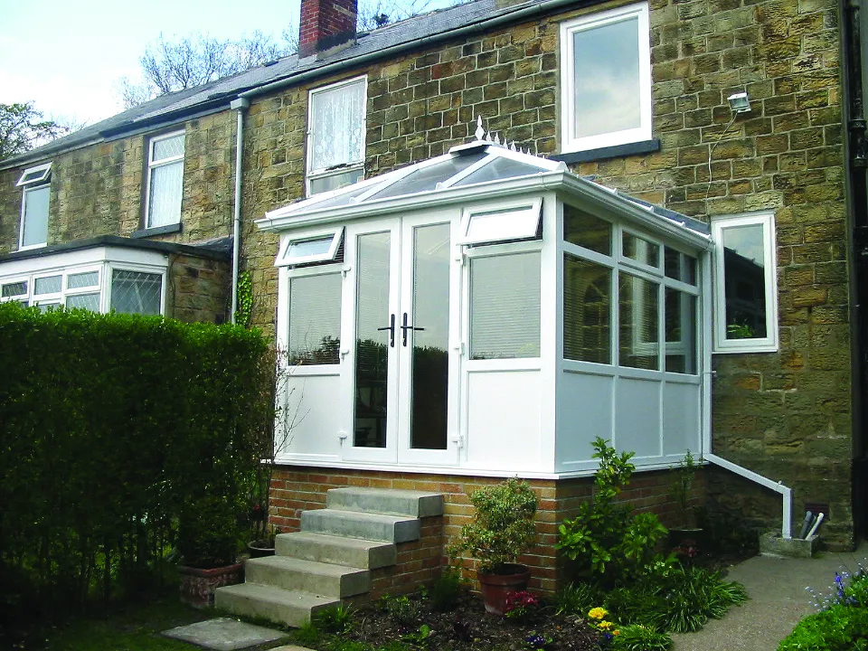 Small white uPVC conservatory built on a brick base with steps leading down to a garden path behind a stone house.