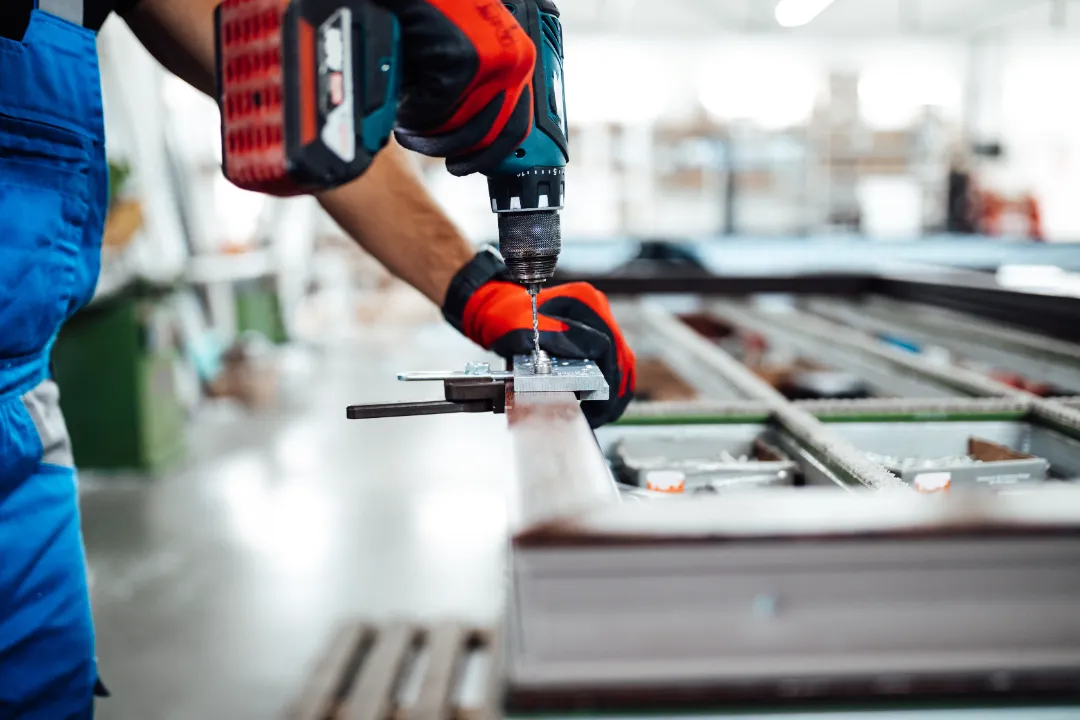 Close-up of a worker in blue overalls drilling precision holes into a window frame using a cordless power drill and mounting jig.