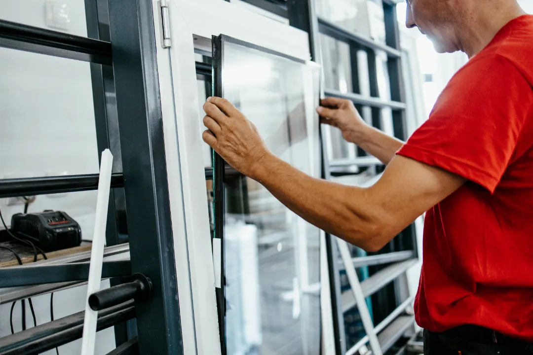 Close-up of a worker in a red t-shirt manually aligning a double-glazed glass unit inside a white window frame structure.