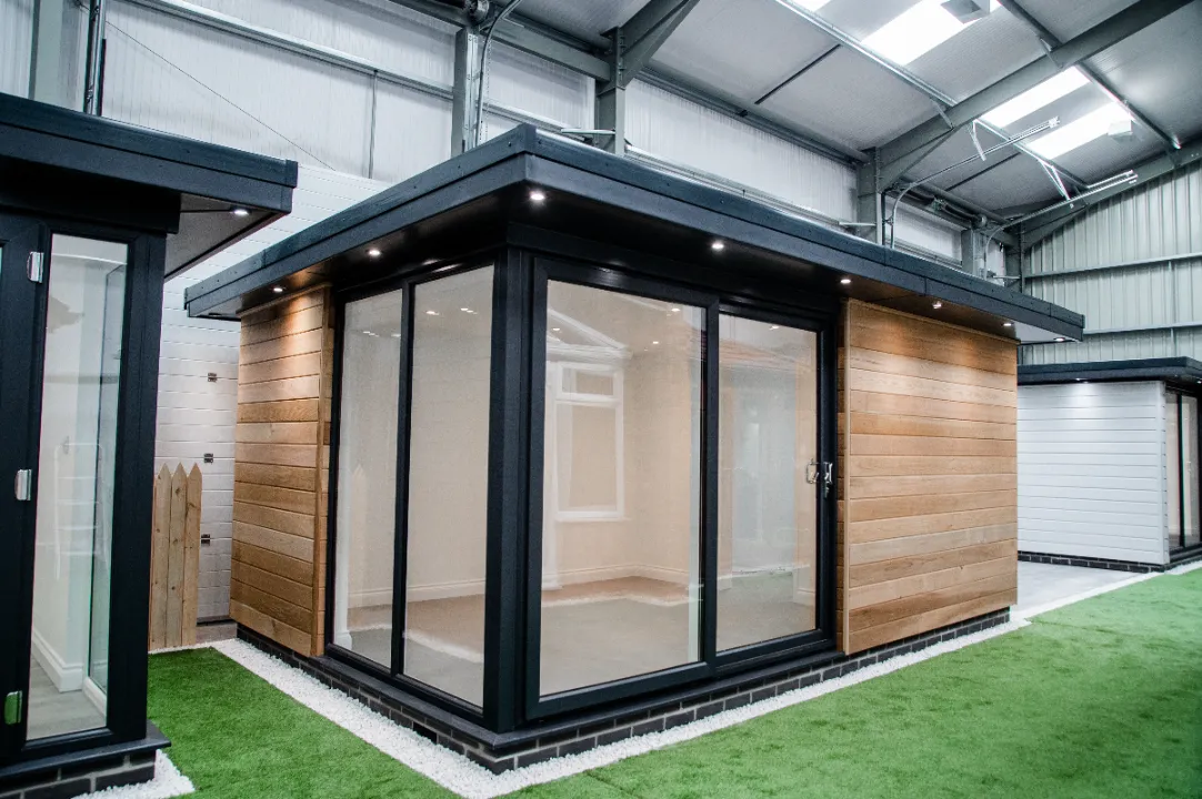 Modern garden room pod with natural wood siding, dark trim, large glass sliding doors, and recessed roof lighting inside a showroom.