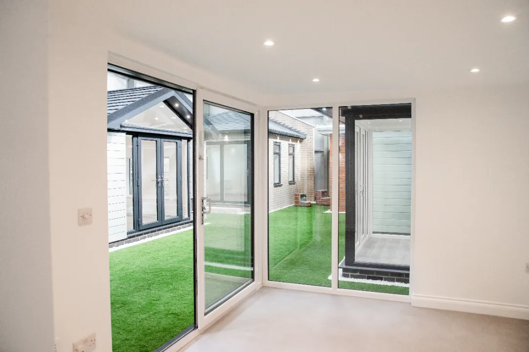 View from inside a white room looking out through floor-to-ceiling glass sliding doors onto a showroom area with artificial turf and display pods.