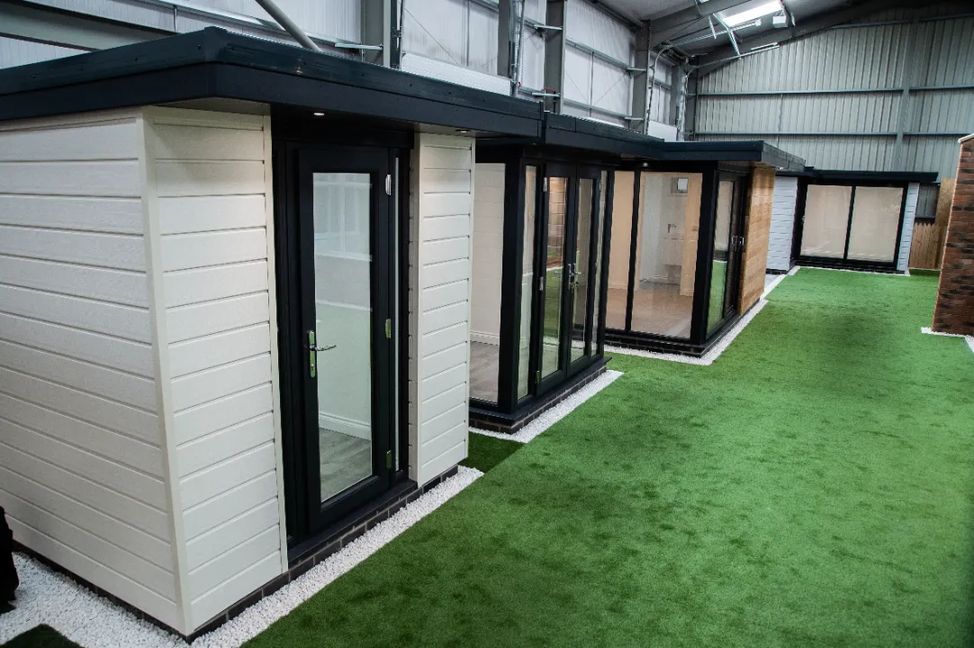 Row of various modern modular garden office pods and outdoor structures displayed inside a large warehouse showroom.