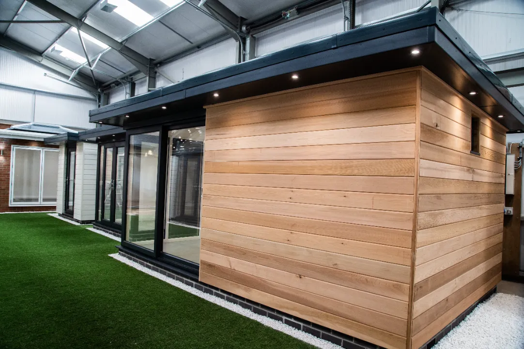 Exterior side view of a modern timber-clad garden room pod with dark overhang lighting set on white gravel and artificial grass.